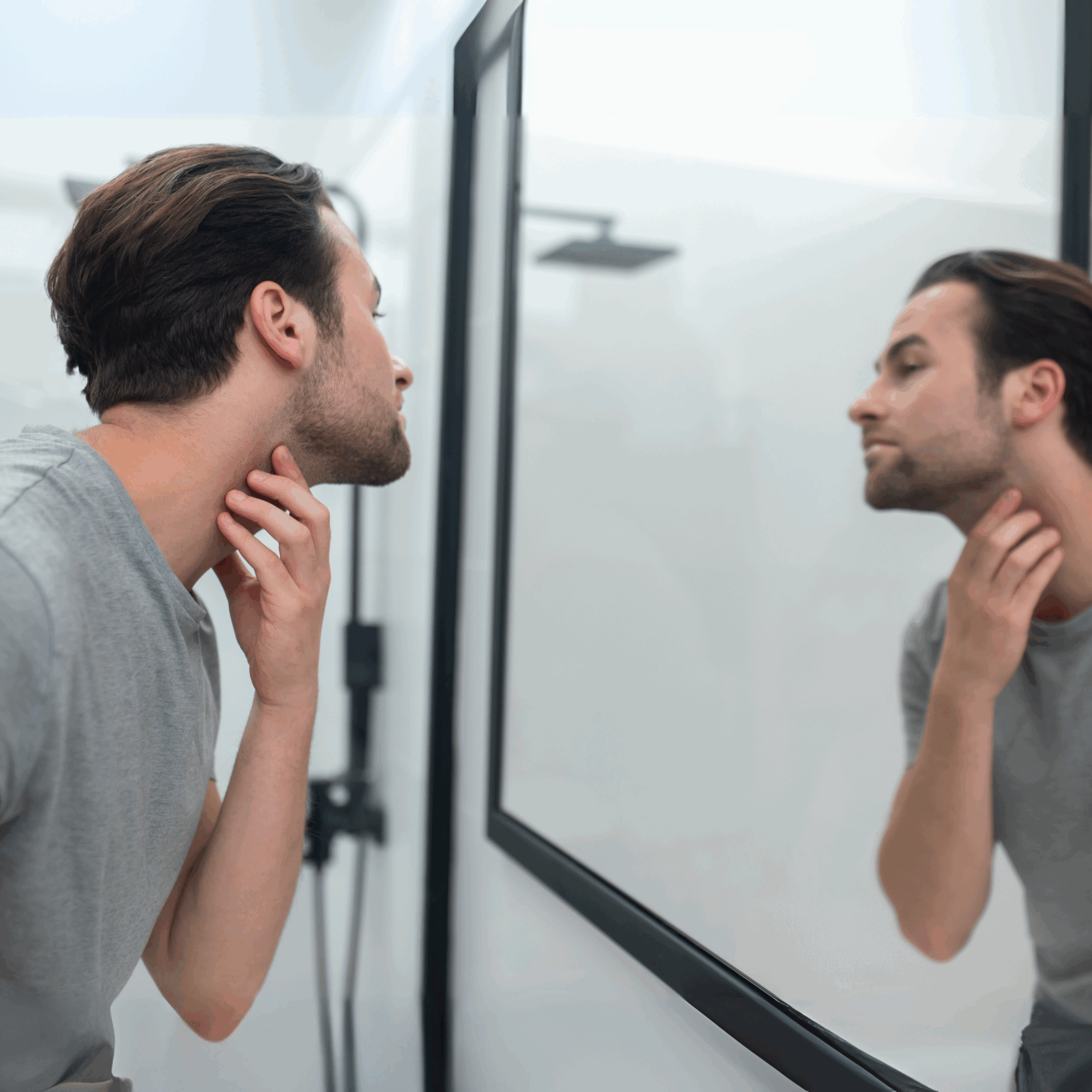 Fiatal férfi néz tükörbe egy fürdőszobában, oldalra fordított fejjel, kezével az állkapcsát vizsgálja, az állvonalát figyeli.