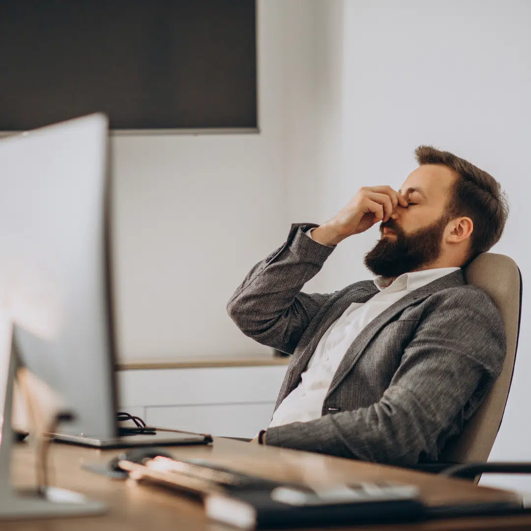 Fáradt férfi ül irodai székben, a szemét dörzsöli egy hosszú munkanap közben – a napközbeni kimerültség tipikus jele.